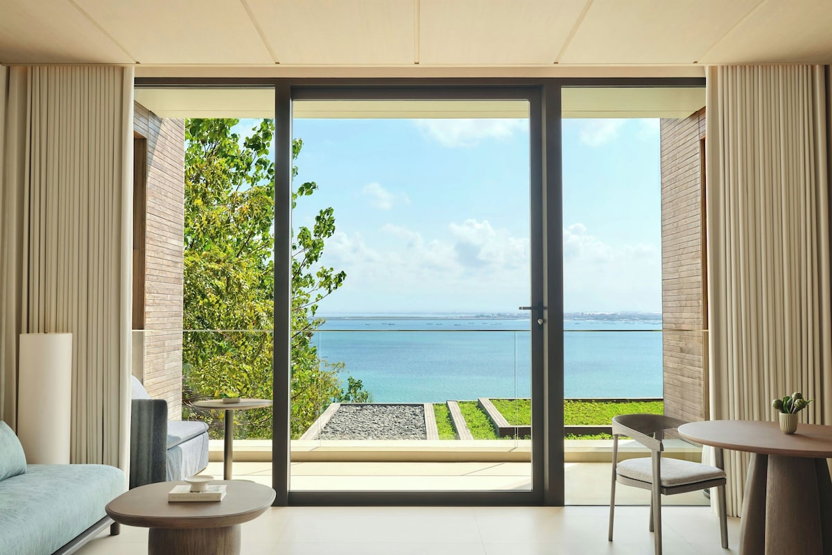 A modern room with a sliding glass door leading to a balcony overlooking a body of water and greenery.