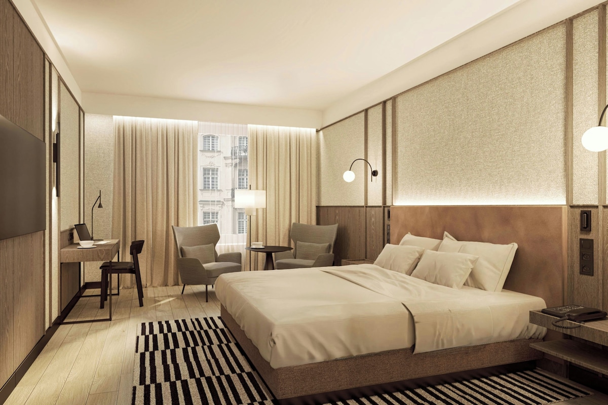 Habitación de hotel con una cama grande, un escritorio con una computadora portátil, dos sillas, una mesita y vistas a edificios a través de la ventana.