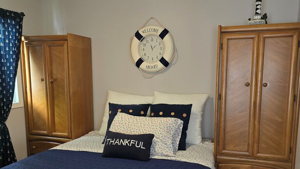 A bedroom with a bed, a wooden wardrobe, a wall clock, and a window with curtains. - Welcome to the Lake House.  A cozy 2 Bedroom Cottage just steps from the Beach. (Central Elgin)