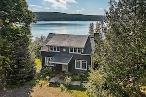 Ein zweigeschossiges Haus mit dunklem Dach und weißem Zierstreifen, umgeben von dichten Bäumen und einem See im Hintergrund.