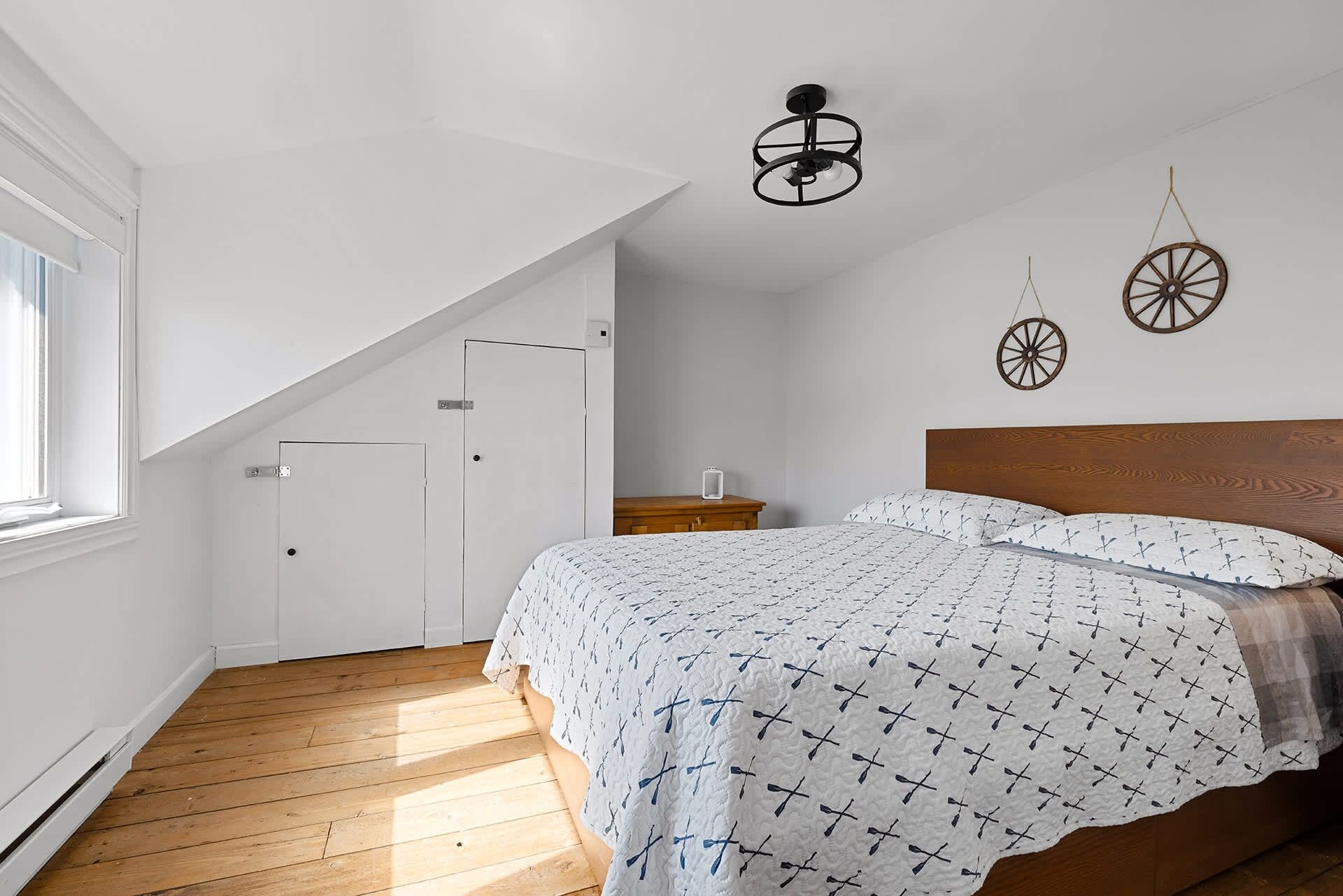 A bedroom with a sloped ceiling, a wooden bed, a bedside table, and a wall-mounted light fixture.