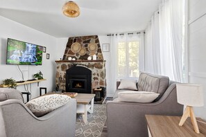 A living room with a stone fireplace, grey sofas, a television, and a large window with curtains. - Clairière sur le Lac - Mandeville - Lake Waterfront (Mandeville)