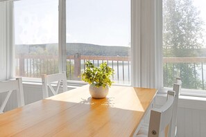 Mesa de jantar de madeira com planta em vaso no centro, cadeiras brancas e vista para um lago através de grandes janelas.
