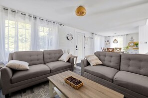 A modern living room with two grey sofas, a wooden coffee table, and a dining area in the background. - Clairière sur le Lac - Mandeville - Lake Waterfront (Mandeville)