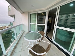A balcony with a glass table and wicker chairs, overlooking a cityscape and a body of water. - Apartment facing Barra da Tijuca beach in the former Sheraton. (Rio de Janeiro)
