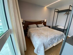 A hotel room with a bed, bedside tables, a hanging light fixture, and a window with curtains. - Apartment facing Barra da Tijuca beach in the former Sheraton. (Rio de Janeiro)