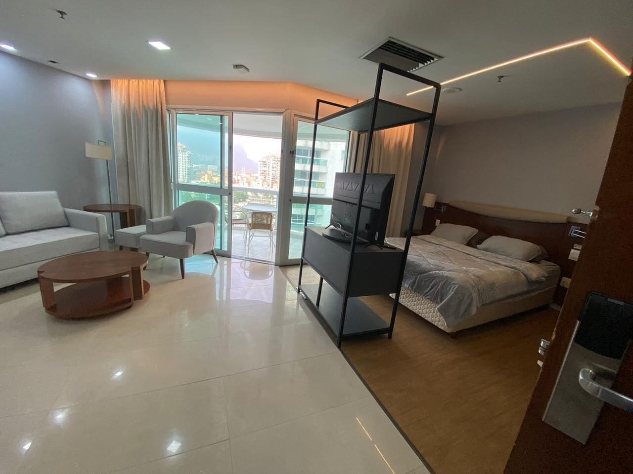 A modern hotel room with a bed, a television, a sofa, a coffee table, and a balcony with a city view.
