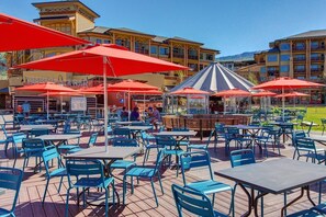 An outdoor patio with tables and chairs, red umbrellas, and a building with balconies in the background. - Luxury Ski In/Out Condo | Pool & Hot tub (Park City)