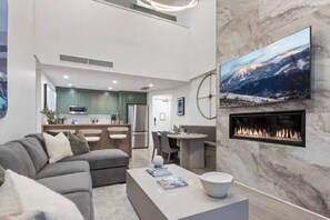 A modern living room with a fireplace, a television mounted on the wall, and a kitchen in the background. - Luxury Ski In/Out Condo | Pool & Hot tub (Park City)