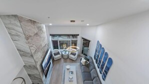 Ein modernes Wohnzimmer mit hoher Decke, einer zentralen Leuchte und einer Sitzecke mit Sofa und Sesseln.