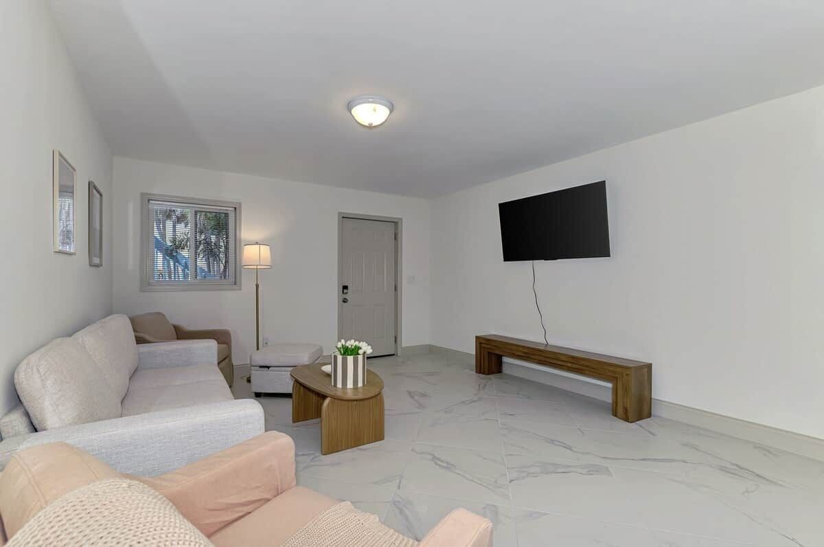 Un salon comprenant un canapé, une table basse, un banc, un téléviseur fixé au mur et une porte.