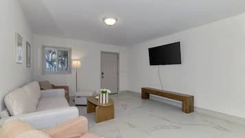 Un salon comprenant un canapé, une table basse, un banc, un téléviseur fixé au mur et une porte.