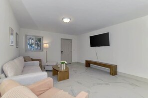 A living room with a sofa, a coffee table, a bench, a TV mounted on the wall, and a door. - Stylish Coastal 1BR | Gulf & Bay Access |Sleeps 4 (Manasota Key)