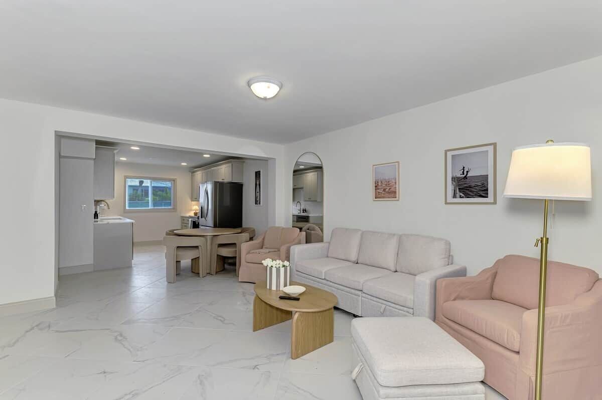 A modern living room with a sofa, a coffee table, and a TV. There is a kitchen in the background with a refrigerator and a dining area.