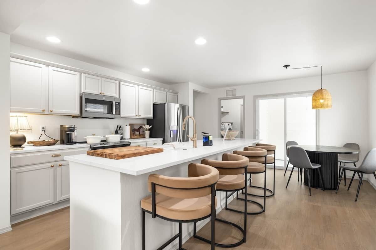A modern kitchen with white cabinetry, a central island with stools, a dining area with a table and chairs, and a large window providing natural light.