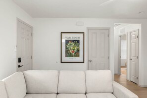 Ein farblich neutral gestaltetes Wohnzimmer mit einem gerahmten Kunstwerk an der Wand, einem Sofa und einer Tür zu einem weiteren Raum.