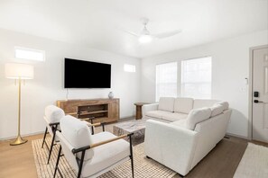 Ein Wohnzimmer mit Flachbildfernseher, einer Couch, zwei Sesseln, einem Couchtisch und einer Stehlampe.