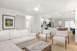 Ein modernes Wohnzimmer mit einer Couch, Sesseln und einem Couchtisch. Im Hintergrund ist eine Küche mit Schränken und Geräten zu sehen.
