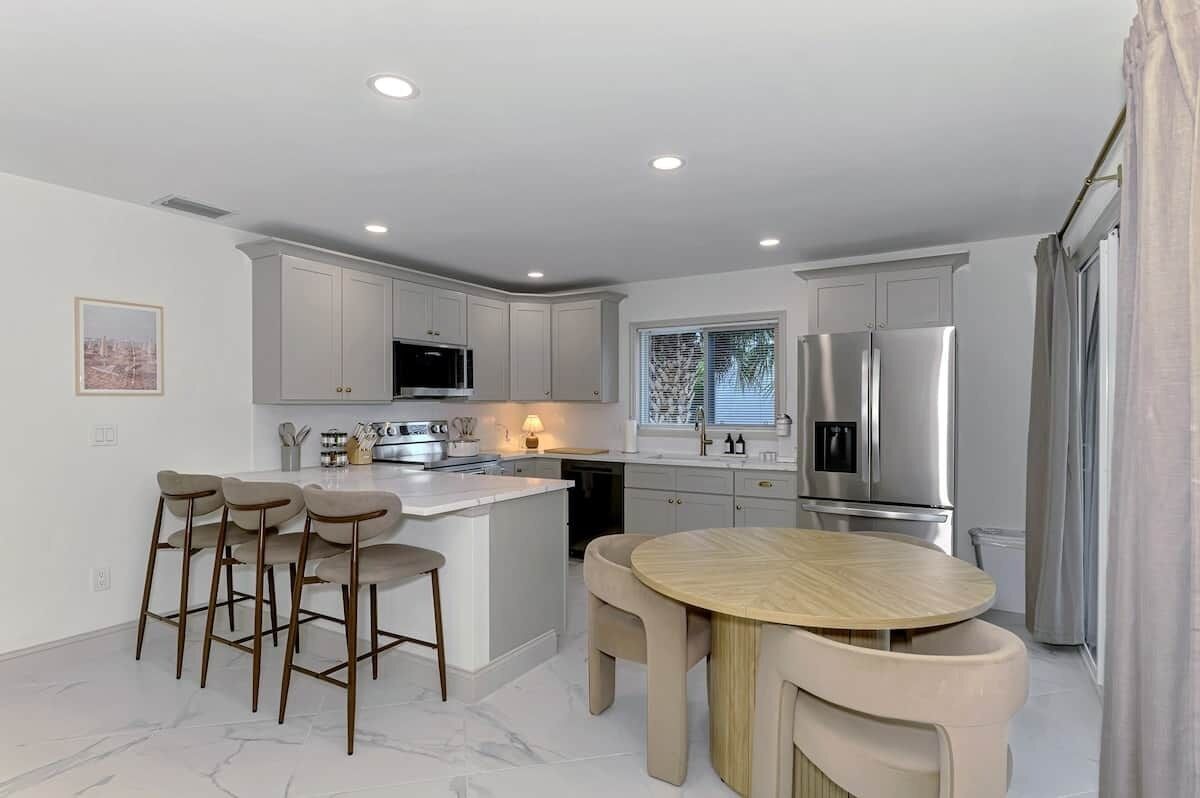 Cocina moderna con gabinetes blancos, electrodomésticos de acero inoxidable, una isla central con taburetes de bar y una mesa de comedor redonda.