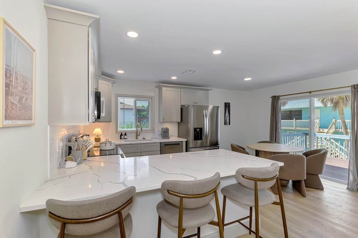 A modern kitchen with a central island, bar stools, and a dining area with a view of a balcony.