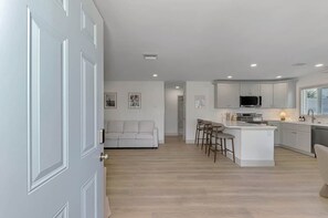 A modern kitchen with white cabinetry, a central island with bar stools, and a dining area with a sofa. - Coastal 2BR Beach Getaway | Gulf + Bay | Unit B (Manasota Key)