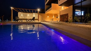 A swimming pool with lounge chairs and a stone wall backdrop, surrounded by a building with multiple balconies and outdoor lighting.