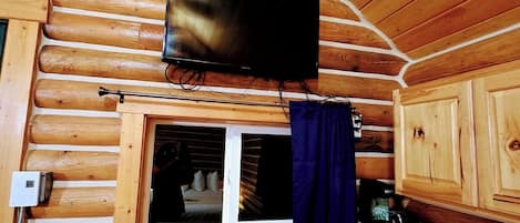 A cabin interior with a television mounted on the wall, a window with curtains, a microwave, and a table with a sign and bottles.