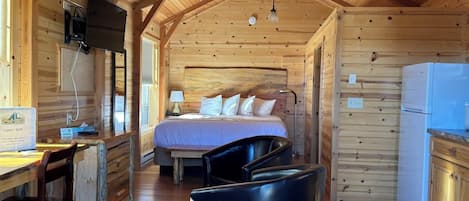 A cabin interior with a bed, a desk, a chair, a refrigerator, and wooden walls.