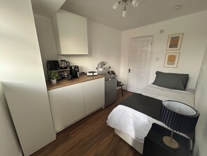 Ein kleines Hotelzimmer mit Küchenzeile, einem Bett mit grauem Kopfteil und einer Nachttischlampe.