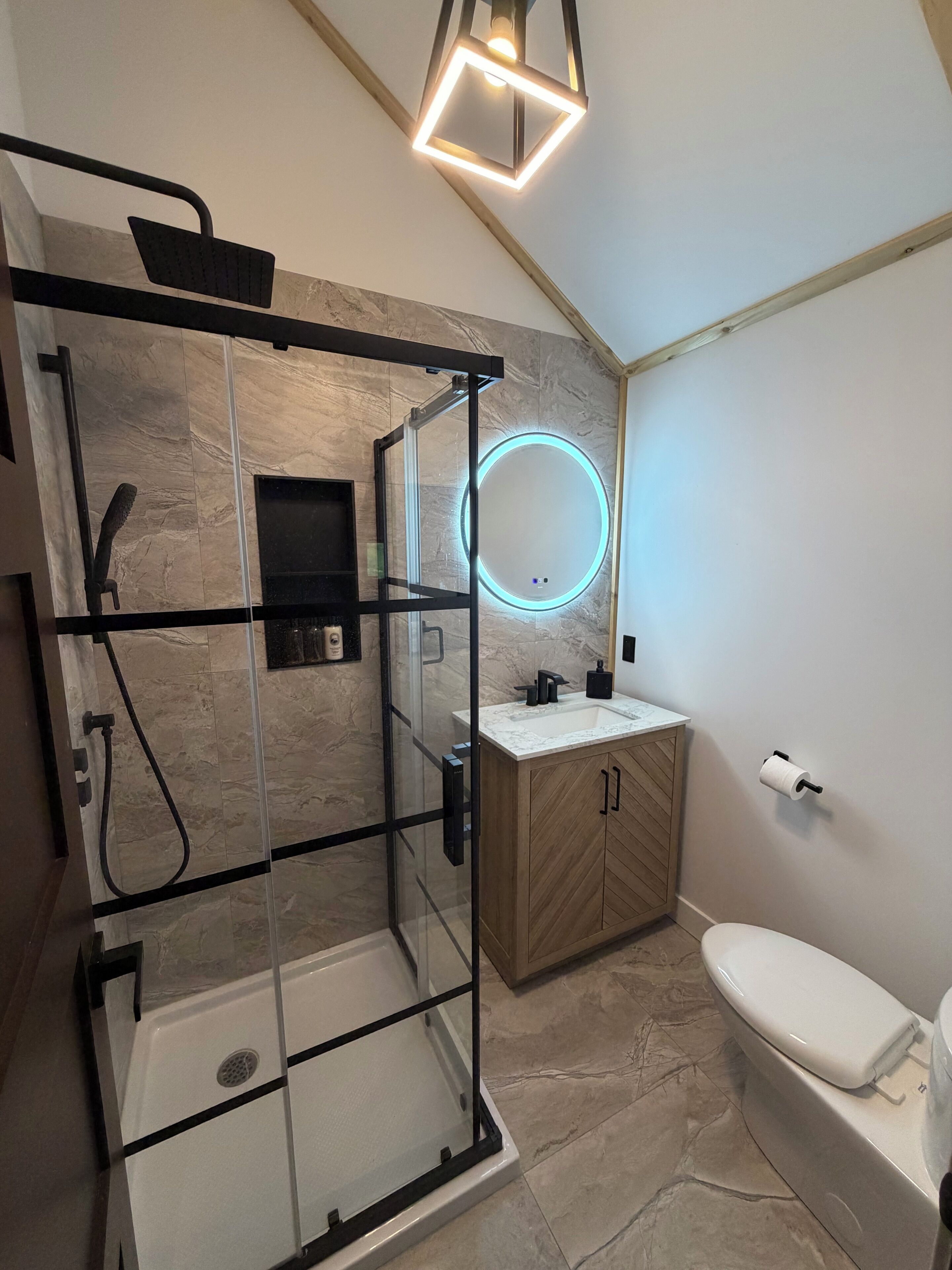 Un baño moderno con ducha envuelta en vidrio, tocador de madera, inodoro y espejo circular con luz integrada en la pared.