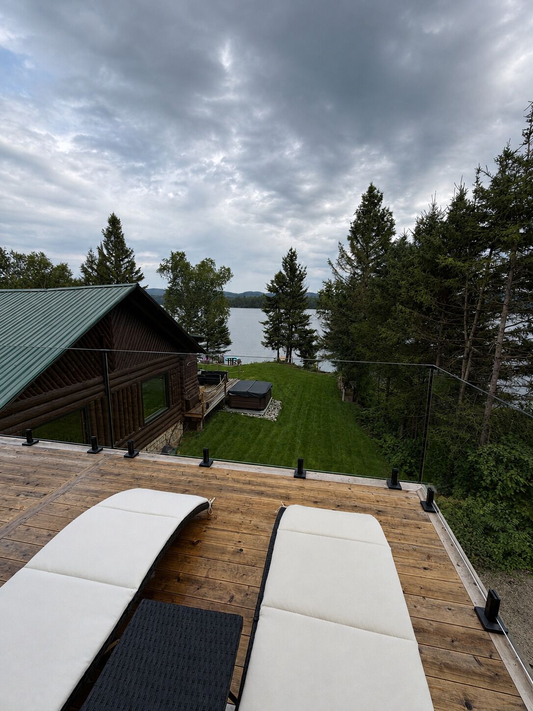 Eine Holzterrasse mit Liegestühlen mit Blick auf einen See, eine Holzhütte und eine Rasenfläche.