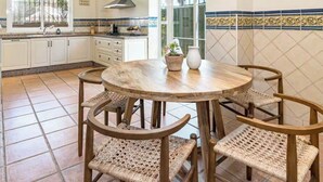 Een keuken met witte kasten, een ronde houten tafel, rieten stoelen en een raam met uitzicht.