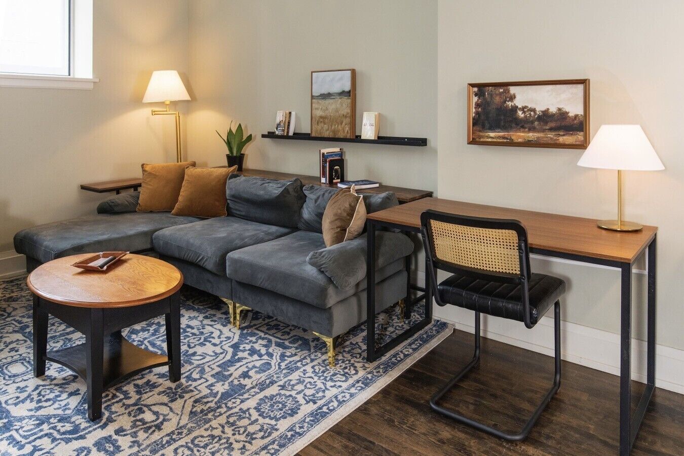 Ein Wohnzimmer mit einem Ecksofa, einem runden Couchtisch, einem Schreibtisch mit Stuhl und zwei Wandleuchten.