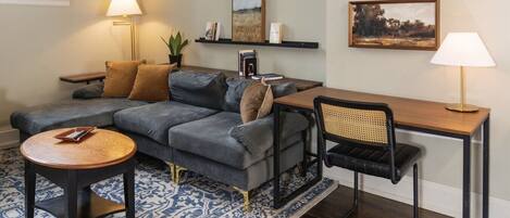 Ein Wohnzimmer mit einem Ecksofa, einem runden Couchtisch, einem Schreibtisch mit Stuhl und zwei Wandleuchten.