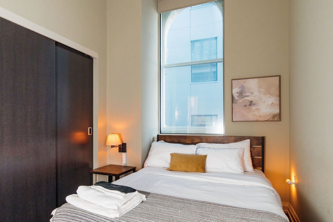Habitación de hotel con cama, mesita de noche, lámpara, ventana y un cuadro en la pared.