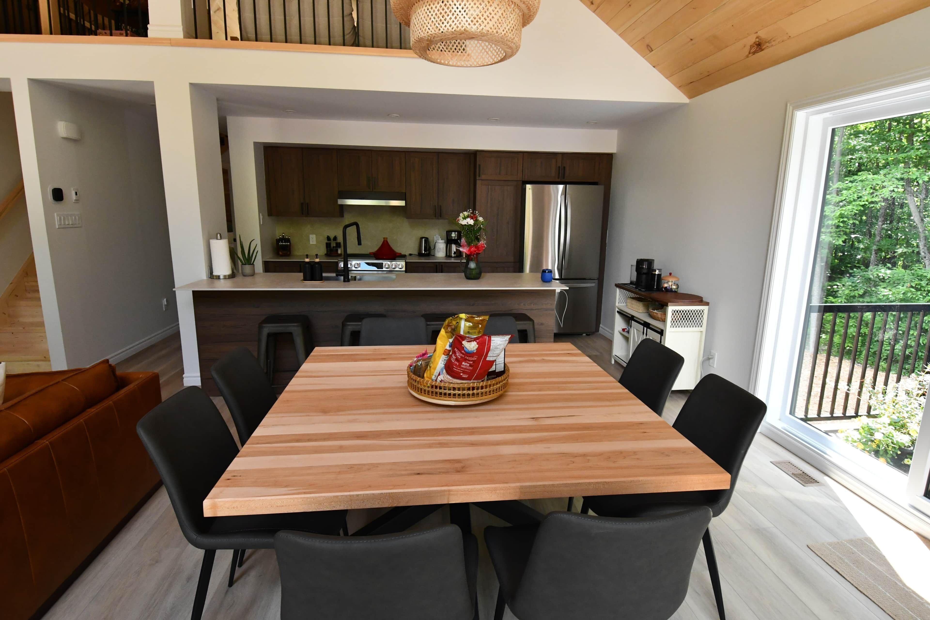 A modern kitchen with wooden cabinets, a stainless steel refrigerator, and a dining area with a wooden table and black chairs.