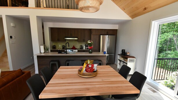A modern kitchen with wooden cabinets, a stainless steel refrigerator, and a dining area with a wooden table and black chairs. - Kozy KanataTremblant (261) (Amherst)