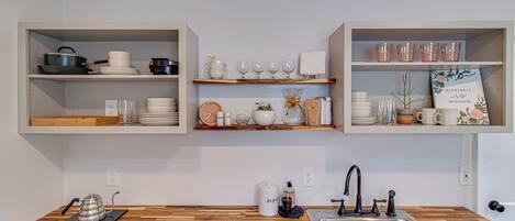 Une cuisine dotée de plans de travail en bois, d’un évier encastré et d’étagères au-dessus, où sont rangés les ustensiles de cuisine et des objets décoratifs.