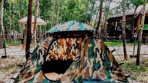 A camouflage tent in a forested area with a wooden structure in the background.