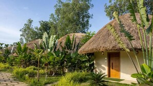 A thatched-roof structure with a brick door, surrounded by lush greenery and a paved pathway.