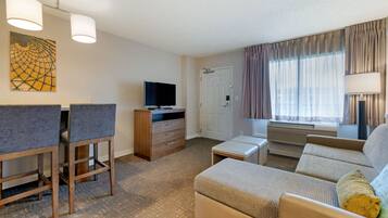 A hotel room with a sofa, a television, a desk with a chair, a door, and a window with curtains.