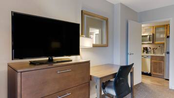 A hotel room with a flat-screen TV on a wooden dresser, a desk with a chair, and a kitchen area with cabinets and appliances.