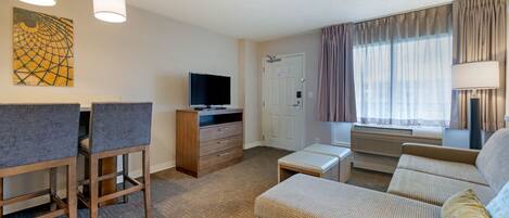 Habitación de hotel con sofá, televisor, escritorio con silla, puerta y ventana con cortinas.