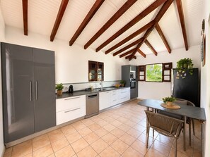 A kitchen with a tiled floor, wooden beams, a dining area with a table and chairs, and a refrigerator with magnets. - Villa Horizon Bleu (SAINTE-LUCE)