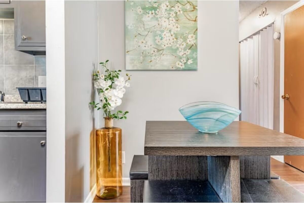 A modern kitchen with grey cabinets, a wooden dining table, a vase with flowers, and a wall-mounted painting.