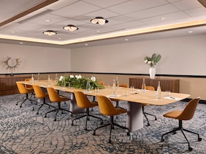 Un salón de conferencias con una larga mesa, varias sillas y una planta decorativa.