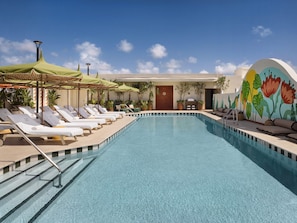 Una piscina con sillones de descanso y sombrillas, una puerta y un muro con un mural floral.