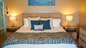 A neatly made bed with a patterned bedspread, two bedside lamps, and a painting of a coastal scene above the bed. - Large Waterfront Retreat with Deep Water Dock! (St Augustine)