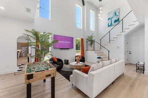 A spacious living area with a foosball table, a large flat-screen TV, a staircase, and a modern kitchen in the background. - Dallas Retreat, Mins to DT, Hot Tub & Big Backyard (Dallas)