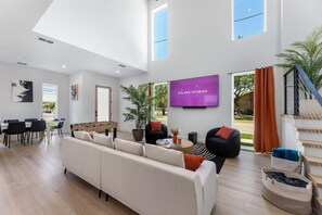 Ein modernes Wohnzimmer mit Sofa, Couchtisch und Fernseher. Es gibt eine Essecke mit Stühlen und einem Tisch. Im Hintergrund ist eine Treppe zu sehen.